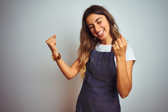 Young beautiful woman wearing apron over grey isolated background very happy and excited doing winner gesture with arms raised, smiling and screaming for success. Celebration concept.
