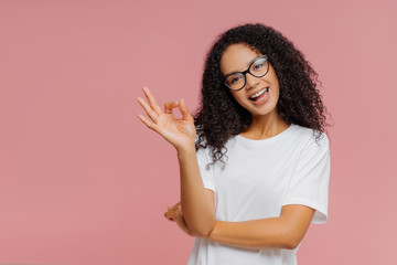 Fototapeta premium Optimistic lovely dark skinned woman makes okay gesture, tilts head, demonstrates approval, agrees with something, has happy expression, wears glasses and casual white t shirt, isolated on pink wall