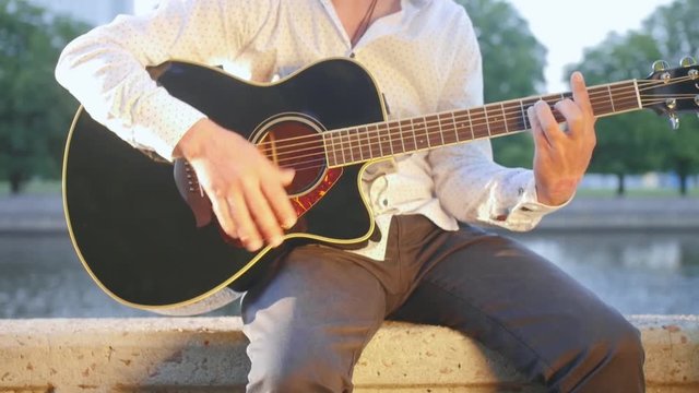 Man sits on a stone boardwalk in the park and plays the guitar. Close up shot.