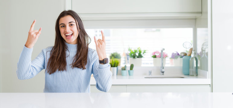 Wide angle picture of beautiful young woman sitting on white table at home shouting with crazy expression doing rock symbol with hands up. Music star. Heavy concept.