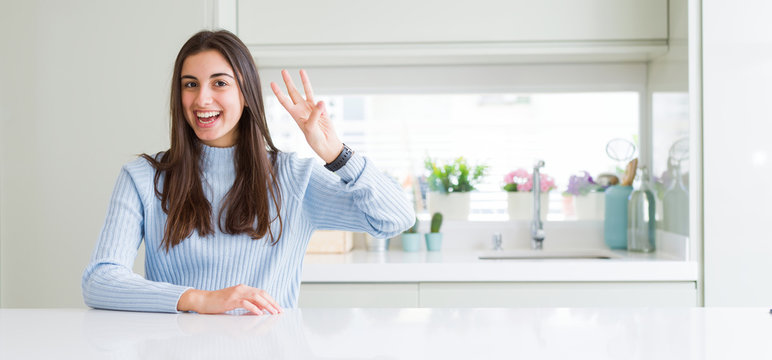 Wide angle picture of beautiful young woman sitting on white table at home showing and pointing up with fingers number three while smiling confident and happy.