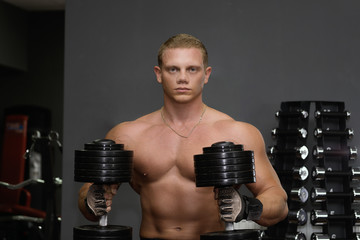 Muscular young man sitting with dumbbells on the bench in the gym and looking into camera. Sport, lifestyle, training and health care concept.