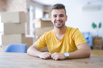 Fototapeta premium Young man sitting on the table with cardboard boxes behind him moving to new home with a happy and cool smile on face. Lucky person.