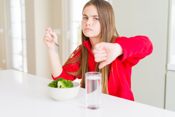 Beautiful young girl kid eating fresh broccoli and drinking water with angry face, negative sign showing dislike with thumbs down, rejection concept