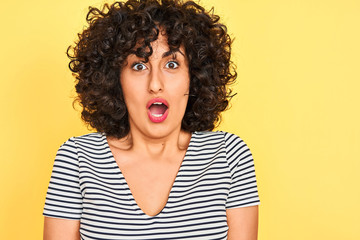 Young arab woman with curly hair wearing striped dress over isolated yellow background scared in shock with a surprise face, afraid and excited with fear expression