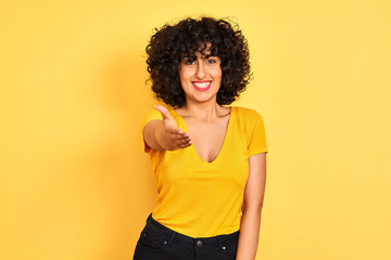 Young arab woman with curly hair wearing t-shirt standing over isolated yellow background smiling friendly offering handshake as greeting and welcoming. Successful business.