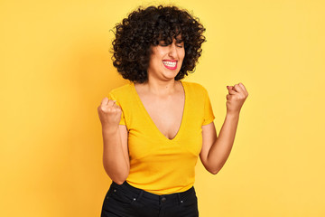Young arab woman with curly hair wearing t-shirt standing over isolated yellow background very happy and excited doing winner gesture with arms raised, smiling and screaming for success. 