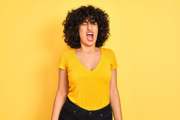 Young arab woman with curly hair wearing t-shirt standing over isolated yellow background angry and mad screaming frustrated and furious, shouting with anger. Rage and aggressive concept.