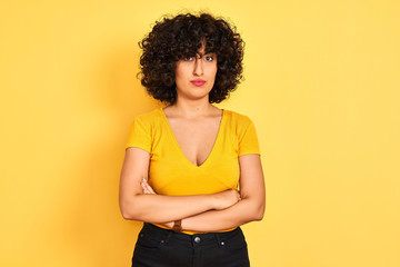 Young arab woman with curly hair wearing t-shirt standing over isolated yellow background skeptic and nervous, disapproving expression on face with crossed arms. Negative person.