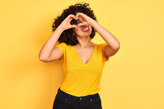 Young arab woman with curly hair wearing t-shirt standing over isolated yellow background Doing heart shape with hand and fingers smiling looking through sign