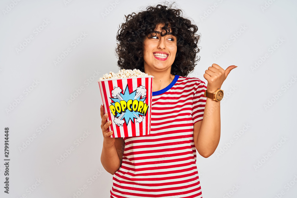 Poster Young arab woman with curly hair holding pack of popcorns over isolated white background pointing and showing with thumb up to the side with happy face smiling