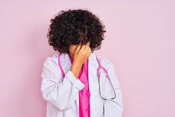 Young arab doctor woman with curly hair wearing stethoscope over isolated pink background tired rubbing nose and eyes feeling fatigue and headache. Stress and frustration concept.