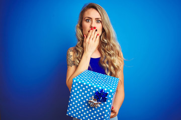 Young beautiful woman holding birthday present over blue isolated background cover mouth with hand shocked with shame for mistake, expression of fear, scared in silence, secret concept