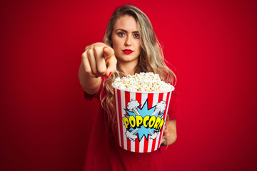 Young beautiful woman wearing t-shirt eating popcorn over red isolated background pointing with finger to the camera and to you, hand sign, positive and confident gesture from the front