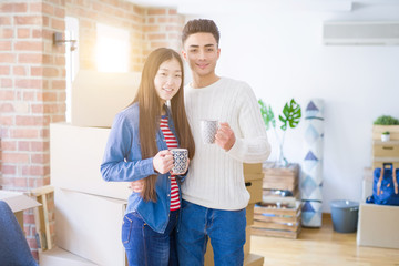 Young asian couple drinking a cup of coffee at new home, smiling happy around cardboard boxes from moving