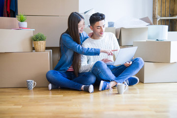 Young asian couple sitting on the floor of new house arround cardboard boxes using laptop and drinking a cup of coffee