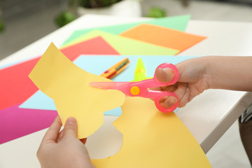 Child cutting out paper heart with plastic scissors at table, closeup