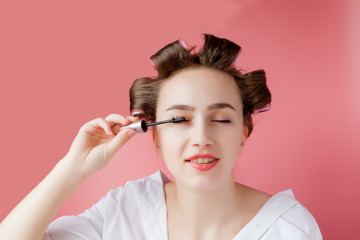 Fototapeta premium nice cheerful young the girl in the curler paints an eyebrow on pink background