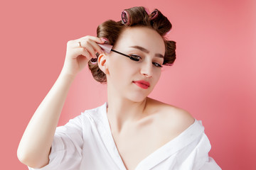 Fototapeta premium nice cheerful young the girl in the curler paints an eyebrow on pink background