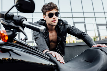 selective focus of handsome man in sunglasses and black leather jacket leaning on motorcycle and looking forward