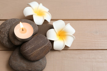 Pile of spa stones with lit candle and flowers on wooden background, space for text
