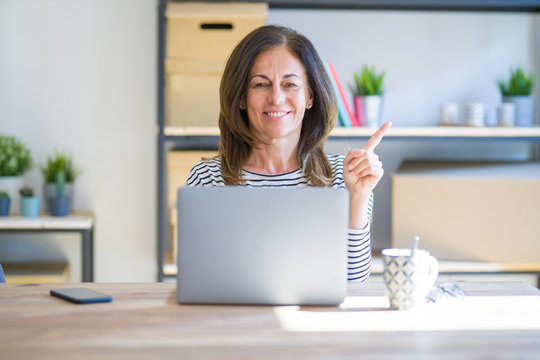Middle age senior woman sitting at the table at home working using computer laptop with a big smile on face, pointing with hand and finger to the side looking at the camera.