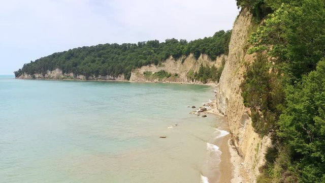 Magnificent sea bay with turquoise water surrounded by impregnable rocks covered with lush forest. Steep cliff off coast. Small soft waves with foam concerning coastal rocks. Aerial takeoff view