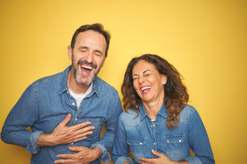 Beautiful middle age couple together standing over isolated yellow background smiling and laughing hard out loud because funny crazy joke with hands on body.