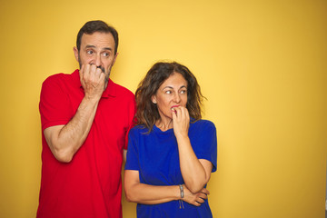 Fototapeta premium Beautiful middle age couple together standing over isolated yellow background looking stressed and nervous with hands on mouth biting nails. Anxiety problem.