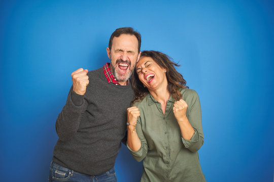Beautiful middle age couple together standing over isolated blue background very happy and excited doing winner gesture with arms raised, smiling and screaming for success. Celebration concept.