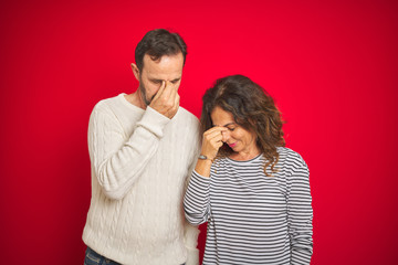 Naklejka premium Beautiful middle age couple wearing winter sweater over isolated red background tired rubbing nose and eyes feeling fatigue and headache. Stress and frustration concept.