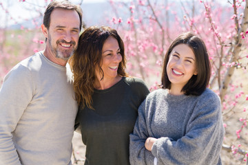 Obraz premium Beautiful family of three smiling cheerful and hugging on peach garden with pink petals enjoying sunny day of spring