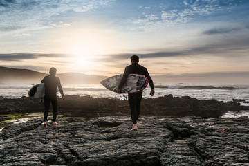 Fototapeta premium two male surfers on rocks with their surfboard on their way to get into the water to surf at sunset