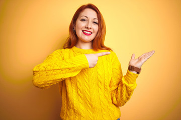 Beautiful redhead woman wearing winter sweater standing over isolated yellow background amazed and smiling to the camera while presenting with hand and pointing with finger.