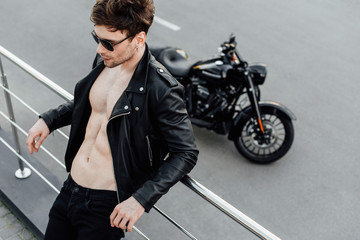 selective focus of man in leather jacket with naked torso leaning on metal fence standing not far from motorcycle