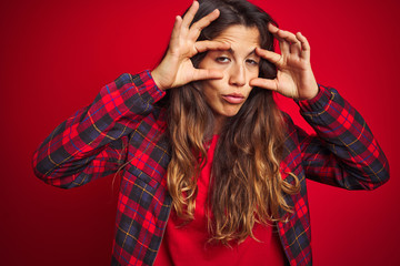 Young beautiful woman wearing casual jacket standing over red isolated background doing ok gesture like binoculars sticking tongue out, eyes looking through fingers. Crazy expression.