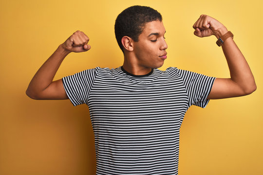 Young handsome arab man wearing navy striped t-shirt over isolated yellow background showing arms muscles smiling proud. Fitness concept.