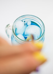 close up woman hand with yellow nail polish holding reusable drinking straw from retro mason glass jar of cold blue fresh non-alcoholic water or cocktail with ice cubs and on light background. 