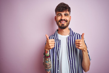 Young man with tattoo wearing striped shirt standing over isolated pink background success sign doing positive gesture with hand, thumbs up smiling and happy. Cheerful expression and winner gesture.