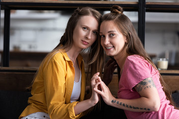 two smiling lesbians touching hands and looking at camera in living room