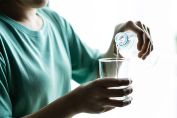 Woman pouring a mineral drinking water from a plastic water bottle into glass close up.  Woman pouring mineral water into glass near the window. Concept of health care and good health life style.