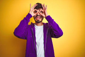 Young man with tattoo wearing sport purple sweatshirt over isolated yellow background doing ok gesture like binoculars sticking tongue out, eyes looking through fingers. Crazy expression.