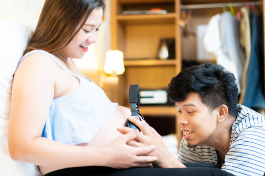 Asian pregnant woman and her hasband putting a wireless headphones on wife's belly, using music for baby revolutionary.