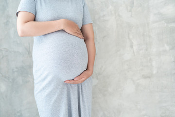Asian pregnant woman in grey prenant dress holds hands on her big belly close up. Concept of healthy pregnant woman, prenant woman's portrait on concrete wall with copy space.