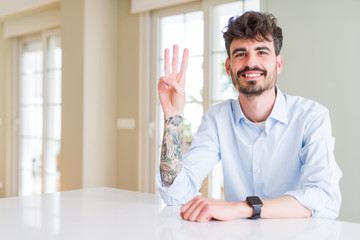 Young businesss man sitting on white table showing and pointing up with fingers number three while smiling confident and happy.