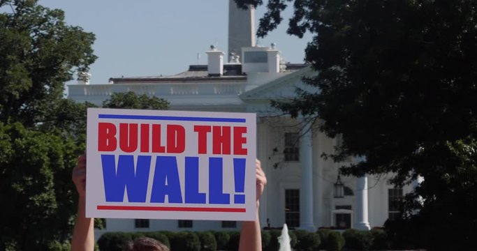 A man holds a Build the Wall protest sign in front of the White House on a sunny summer day.  	