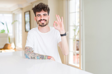 Young man wearing casual t-shirt sitting on white table smiling positive doing ok sign with hand and fingers. Successful expression.