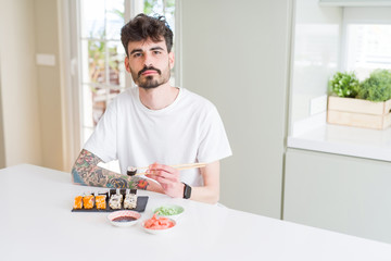 Young man eating asian sushi from home delivery with a confident expression on smart face thinking serious