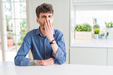 Young man wearing casual shirt sitting on white table bored yawning tired covering mouth with hand. Restless and sleepiness.