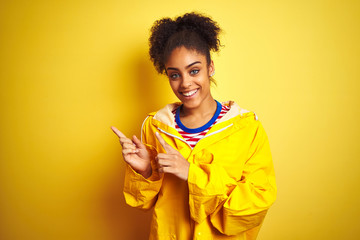 Young african american woman wearing rain coat over isolated yellow background smiling and looking at the camera pointing with two hands and fingers to the side.